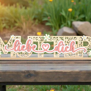 May include: A wooden sign with the German words "Mutter, liebe dich" in a decorative font, with a pink and white accent. The sign is adorned with small white daisies and is set on a wooden table.