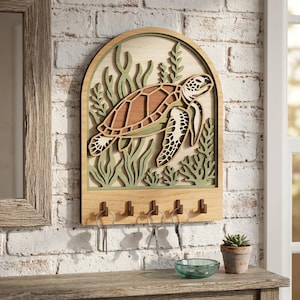 Puede incluir: Llavero de madera con un diseño en capas de una tortuga marina nadando entre algas. El llavero tiene un acabado de madera natural con detalles en verde y marrón. Incluye cinco ganchos para llaves.