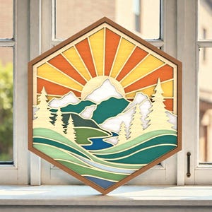 May include: A wooden, multi-layered wall art piece depicting a mountain landscape with a rising sun. The artwork features a hexagonal frame and uses shades of yellow, orange, green, and blue to create depth and dimension. The piece is displayed on a white windowsill.
