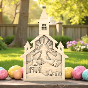 Puede incluir: Una decoración de madera con forma de iglesia con intrincados detalles recortados que representan una escena de la natividad, cruces y un sol radiante. La iglesia está colocada sobre una superficie de madera con coloridos huevos de Pascua delante. El fondo presenta un césped verde y una valla de madera.