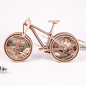 Pode incluir: Uma escultura de madeira em camadas com formato de bicicleta de montanha. As rodas apresentam recortes intrincados que retratam um ciclista numa paisagem montanhosa com árvores, rochas e um sol, em tons de madeira clara e escura.