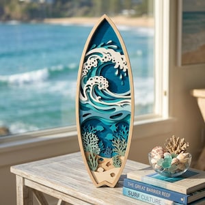 May include: A wooden surfboard-shaped wall art piece featuring a layered ocean scene. The design includes a large wave, coral, and fish, with a natural wood frame. The artwork is displayed on a wooden table near a window overlooking the ocean.