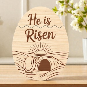 May include: Wooden Easter egg decoration with the text "He is Risen". The design features a sunrise over a tomb. The egg is made of light-colored wood and is suitable for Easter decor.