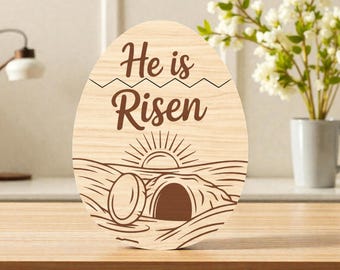 May include: Wooden Easter egg decoration with the text "He is Risen". The design features a sunrise over a tomb. The egg is made of light-colored wood and is suitable for Easter decor.