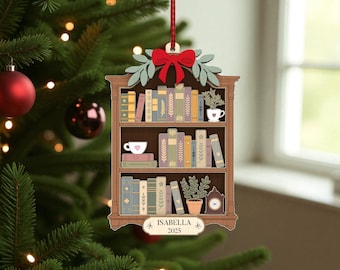 May include: Wooden Christmas tree ornament shaped like a bookshelf, with a red bow and green leaves. The ornament depicts a bookshelf with books, a teacup, and a small clock. The name "Isabella" and the year "2025" are displayed.