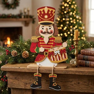 May include: A wooden nutcracker figure dressed in a red uniform with gold epaulets, a tall hat, and a mustache. The figure is playing a drum with red and white stripes and is attached to dangling legs with black boots that hang off the edge.