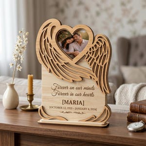 Könnte beinhalten: Gedenk-Fotorahmen aus Holz mit Engelsflügeln und einem herzförmigen Ausschnitt. Der Rahmen enthält ein Foto eines Paares. Der Text lautet "Forever on our minds, Forever in our hearts" mit einem Namen und Daten. Auf einer Holzoberfläche.