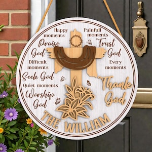 May include: A round wooden sign with a white background and a brown border. The sign features a wooden cross, flowers, and the words "Praise God," "Thank God," and "The William." The sign is hanging on a door.