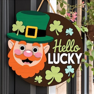 May include: A round wooden St. Patrick's Day door sign featuring a leprechaun face with a green hat, orange beard, and pink cheeks. The sign says "Hello LUCKY" with shamrocks in green and light blue. A rope is attached for hanging.