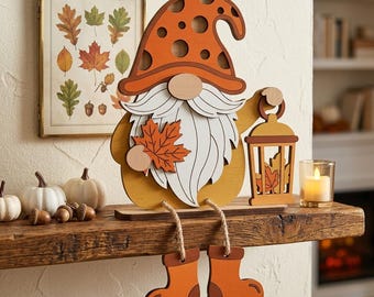 May include: A layered wooden gnome decoration featuring an orange polka-dot hat, a white beard, and a yellow body. The gnome holds a maple leaf and a lantern, with dangling legs wearing orange boots, sitting on a wooden shelf with small pumpkins.
