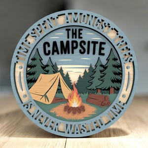 Könnte beinhalten: Runder Holzschild mit einer Camping-Szene. Das Schild zeigt ein Zelt, ein Lagerfeuer und Bäume, mit den Worten "THE CAMPSITE" und "TIME SPENT AMONGST TREES IS NEVER WASTED TIME" in einem kreisförmigen Design. Das Schild ist in Blau-, Grün- und Brauntönen bemalt.