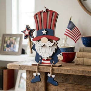 May include: Wooden patriotic gnome figurine in red, white, and blue. The gnome wears a tall hat and jacket, holding a star-shaped wand. The figurine has dangling blue boots and a white beard. An American flag is in the background.