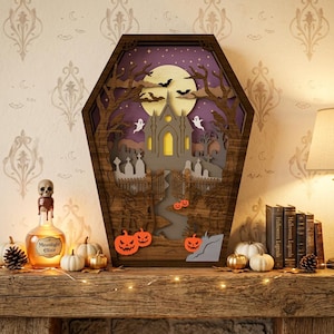 Puede incluir: Una caja de sombra de madera con forma de ataúd que presenta una escena de Halloween en capas con una iglesia embrujada, una luna llena, murciélagos, fantasmas, lápidas y calabazas naranjas. El marco de madera oscura encierra un cielo morado.