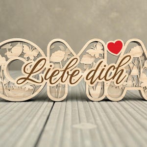 Op de afbeelding: Houten decoratief bord met het woord "OMA" en de zin "Liebe dich" in bruin script. De letters zijn uitgesneden met bloemen- en vlindermotieven. Een rood hart is boven de letter "A" geplaatst.