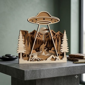 May include: A multi-layered wooden laser-cut display featuring a Bigfoot silhouette walking through a forest with pine trees and mountains. A large UFO hovers above with a beam of light shining down toward the figure, set against a full moon.