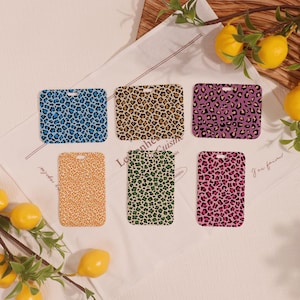 May include: Six rectangular leopard print designs in various colours: blue, brown, purple, orange, green, and pink. Each has a small hole at the top. The items are arranged on a white cloth with lemons.