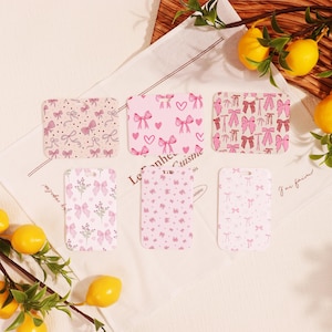 Può includere: Sei sottobicchieri rettangolari con motivi a fiocco rosa. Ogni sottobicchiere presenta un design diverso, tra cui fiocchi, cuori ed elementi floreali. I sottobicchieri sono disposti su un panno bianco con testo e limoni sullo sfondo.