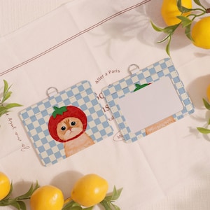 May include: Two light blue and white checkered pattern ID badge holders. One features a cat wearing a tomato hat, the other has a blank window. Both have silver metal rings at the top.