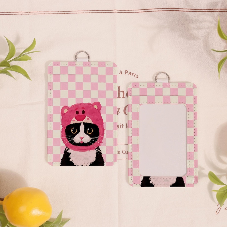 May include: Two pink and white checkered ID badge holders. One features a black and white cat wearing a pink bear hat. The other has a clear window for an ID. Both have a silver ring at the top.