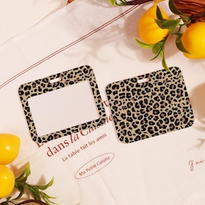 Può includere: Due portabadge rettangolari con motivo leopardato. Uno ha una finestra trasparente, l'altro è pieno. I supporti sono beige con macchie nere. Limoni e un panno bianco con testo francese sullo sfondo.