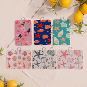 Könnte beinhalten: Sechs rechteckige Dekorationsartikel mit silbernen Schlaufen. Die Artikel weisen verschiedene Muschel- und Seesternmuster in Rosa, Blau und Türkis auf. Die Designs umfassen Korallen, Muscheln und Seesterne. Die Artikel sind auf einer weißen Oberfläche mit Zitronen angeordnet.