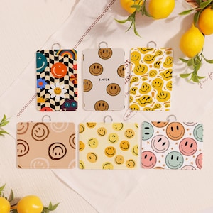 Op de afbeelding: Zes rechthoekige kaarthouders met smiley-gezichten. Patronen omvatten een dambord met een regenboog en smiley-gezicht, en herhaalde smiley-gezichten. Het kleurenpalet omvat geel, bruin en pasteltinten. Elke houder heeft een metalen lus.