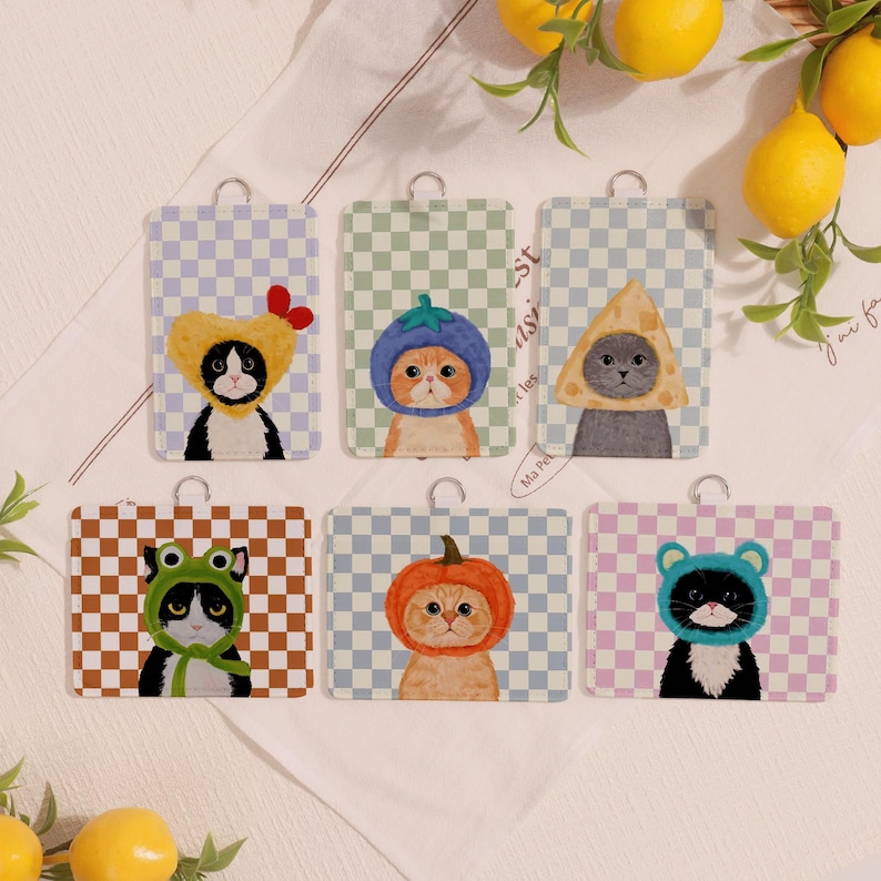 May include: Six rectangular cat-themed tags with silver metal loops. Each tag features a different cat illustration wearing a costume, set against a checkered background. Costumes include a chicken, a blueberry, a cheese wedge, a frog, a pumpkin, and a bear.