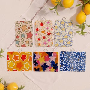 May include: Six rectangular tags with star patterns in various colours. The tags have a silver loop at the top. The star patterns include pastel, red, yellow, and blue stars on different coloured backgrounds. The tags are arranged on a white surface.