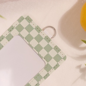 May include: A light green and white checkered pattern ID badge holder with a silver metal ring. The holder has a clear window for displaying an ID card. The edges are stitched with a matching thread.
