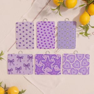 Puede incluir: Seis tarjeteros lavanda con llaveros plateados. Cada tarjetero presenta un patrón diferente: lunares, flores, conejitos, lazos, estrellas y corazones. Los tarjeteros están dispuestos sobre una superficie blanca con limones.