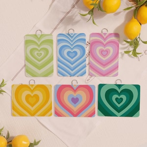 Puede incluir: Seis tarjeteros rectangulares con presillas de metal plateado. Cada tarjetero presenta un diseño de corazón concéntrico en varios esquemas de color: verde, azul, rosa, amarillo, arcoíris y verde oscuro. Los tarjeteros se muestran sobre una superficie blanca con limones.