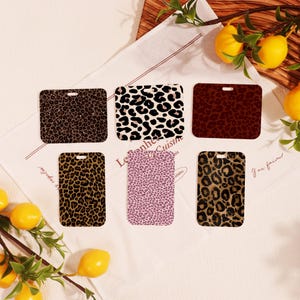 May include: Six rectangular leopard print items in various colours, including brown, black, white, and pink. Each item has a small hole at the top. The items are arranged on a white cloth with lemons and greenery in the background.