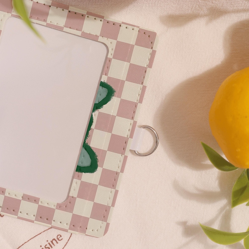 May include: A pink and white checkered card holder with a clear window and a silver ring. The card holder has a green leaf design. A yellow lemon with green leaves is in the background.