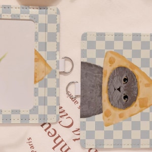 May include: A light blue and white checkered card holder with a cat wearing a cheese hat. The card holder has a clear window and silver metal rings. The cat has big eyes and gray fur.