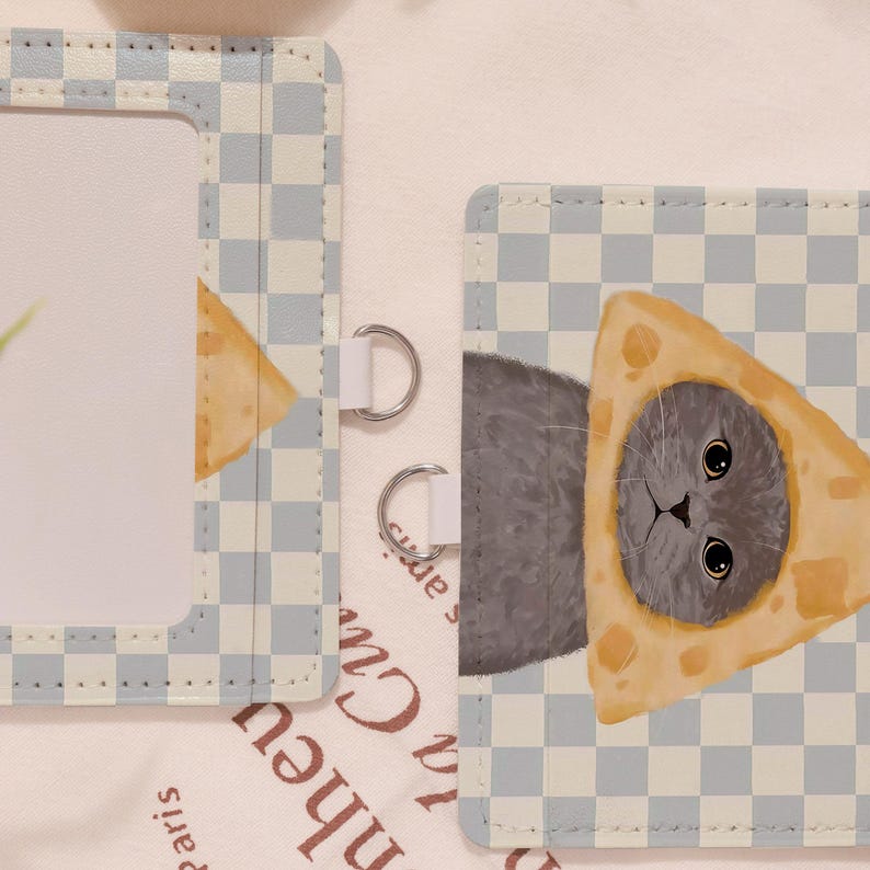 May include: A light blue and white checkered card holder with a cat wearing a cheese hat. The card holder has a clear window and silver metal rings. The cat has big eyes and gray fur.