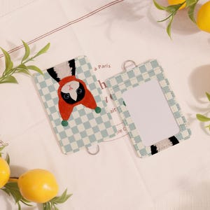 May include: Two light blue and white checkered cardholders. One features a cartoon animal in a red hat, the other has a clear window. Both have silver rings for attaching to a lanyard. The background is white.