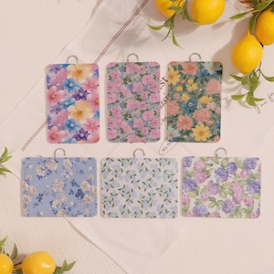 May include: Six rectangular floral-patterned cutting boards with silver hanging loops. The boards feature various flower designs in shades of pink, blue, yellow, and purple. Lemons and greenery are in the background.
