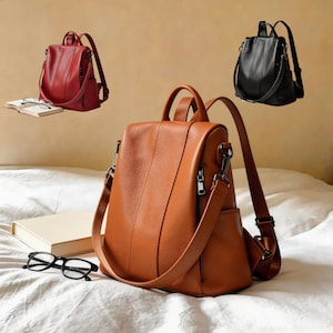 May include: Three leather backpacks in red, black, and brown. The brown backpack is in the foreground, with a zippered pocket and adjustable straps. The red and black backpacks are in the background. A pair of glasses and a book are in the foreground.