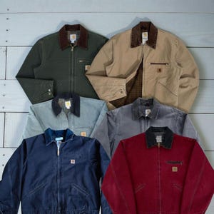 May include: Assortment of Carhartt work jackets in various colors, including green, tan, gray, navy, and red. Each jacket features a zip-up front, a corduroy collar, and a small square logo patch on the chest. The jackets are laid out on a light wood-grain surface.