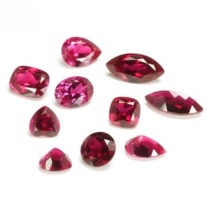 May include: A collection of nine loose faceted gemstones in a vibrant deep pinkish-red hue. The stones are cut into various shapes including pear, marquise, cushion, oval, heart, and round, all featuring precise geometric facets that reflect light.