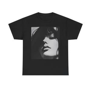 Op de afbeelding: Zwart-witfoto van een vrouwen gezicht. De vrouw heeft donker haar en kijkt naar beneden. De foto is gedrukt op een zwart T-shirt.