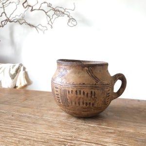 May include: A small, round, tan-coloured earthenware mug with a single handle. The surface features dark brown, hand-painted geometric patterns, including horizontal lines and vertical dashes encircling the body and neck of the vessel.