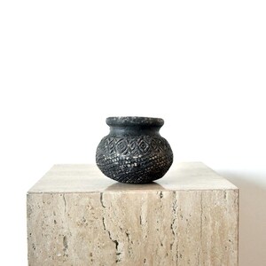 May include: A small, dark grey ceramic pot with a textured surface and a patterned design. The pot sits on a light beige stone block, creating a contrast in textures and colours. The pot's shape is round with a slightly flared rim.