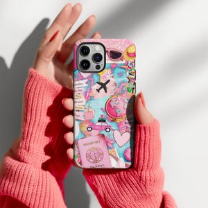 May include: A pink and blue phone case featuring a world map design with travel-themed illustrations. The case includes images of a passport, airplane, car, sunglasses, and a globe. The case is held by a person wearing a pink sweater.