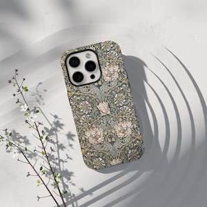 Puede incluir: Una funda de teléfono con un estampado floral en tonos verdes, beige y amarillo. La funda tiene un borde oscuro alrededor de las lentes de la cámara y un acabado mate. El diseño presenta flores y follaje estilizados.