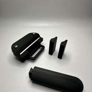 May include: A black 3D-printed tool set consisting of a main housing unit with two silver screws, two small rectangular spacers with curved edges, and a separate elongated handle piece with a textured surface and a triangular connector end.