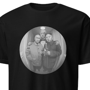 May include: Black t-shirt featuring a grayscale image of three men in a circular frame. The image shows the men in a hallway, with one man in the center and the other two flanking him. The men are wearing jackets and holding bottles.