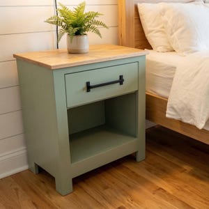 May include: A sage green nightstand with a natural wood top and a black handle. The nightstand has a drawer and an open shelf. A potted fern sits on top of the nightstand. The nightstand is next to a bed with white bedding.