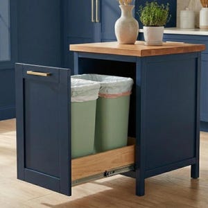 Puede incluir: Isla de cocina azul marino con encimera de madera. La isla cuenta con un cajón extraíble con dos botes de basura verdes en su interior. Un tirador dorado está en la puerta. Un jarrón y una planta en maceta se encuentran en la parte superior de la isla.