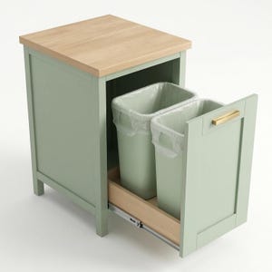 Pode incluir: Um armário de cozinha verde sálvia com um tampo de madeira clara e uma gaveta retrátil. A gaveta revela dois caixotes do lixo de plástico. A porta do armário tem uma pega dourada. Uma solução prática de gestão de resíduos.
